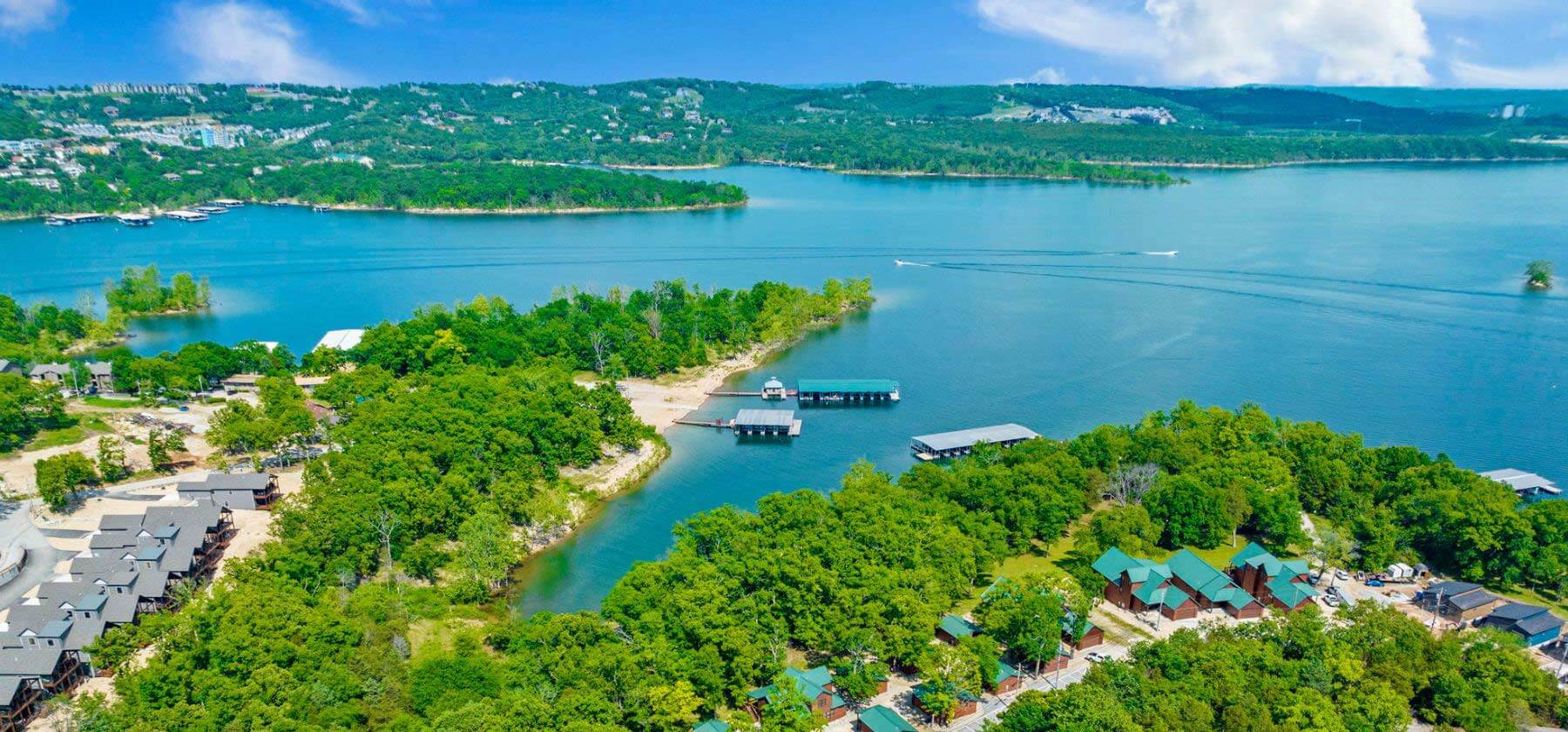 Aerial view of a lush lakeside resort with cabins, docks, and boats on clear waters.