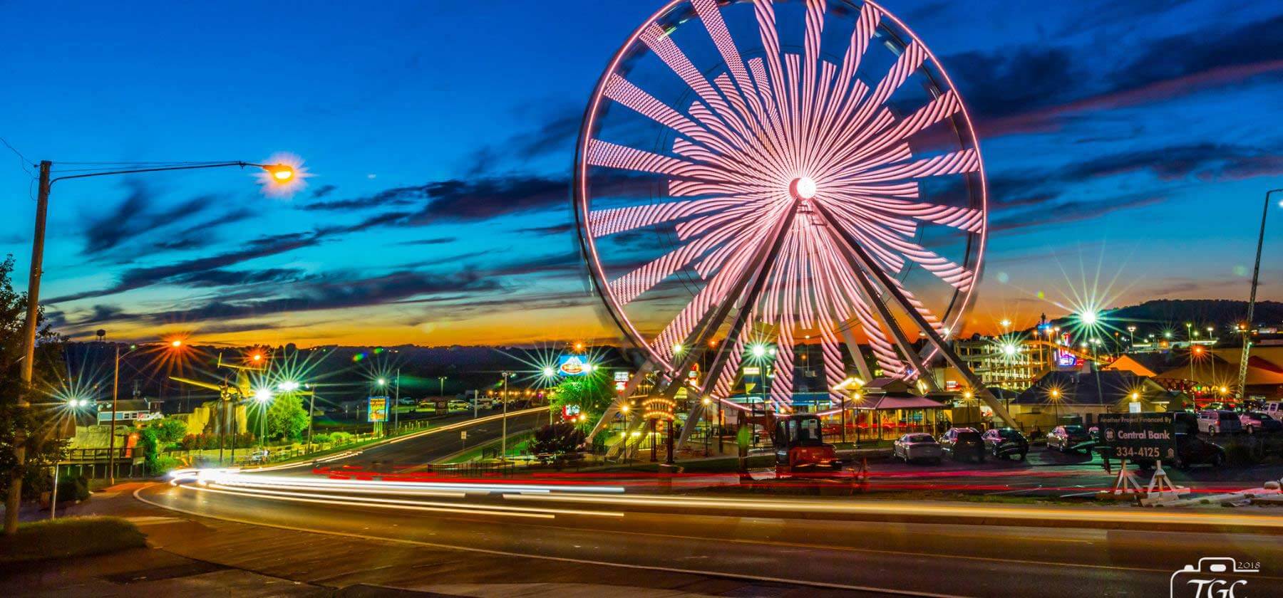 Illuminated Ferris wheel at twilight with long-exposure street lights and vibrant sky.