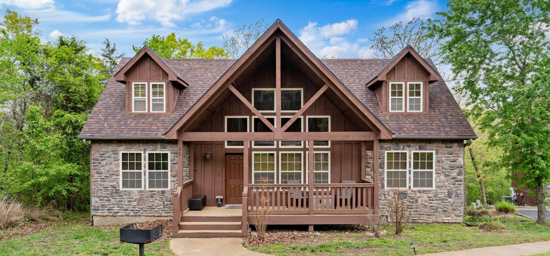 A charming two-story cottage with a stone facade and brown trim, surrounded by greenery.