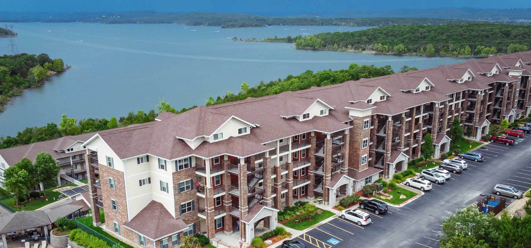 Aerial view of a long residential building complex by a large lake with a forested shoreline.