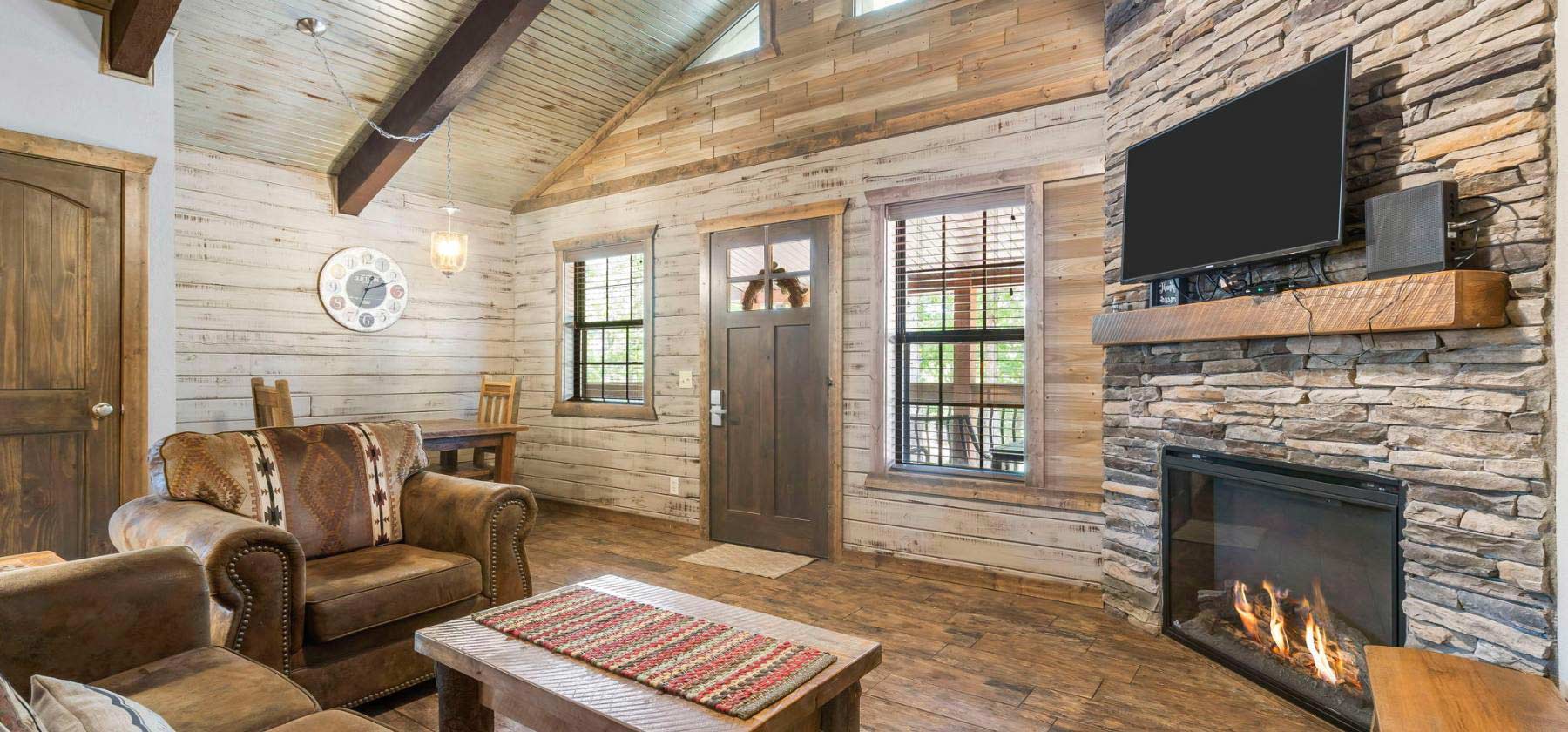 Rustic living room with a fireplace, wooden walls, leather couches, and a mounted TV.