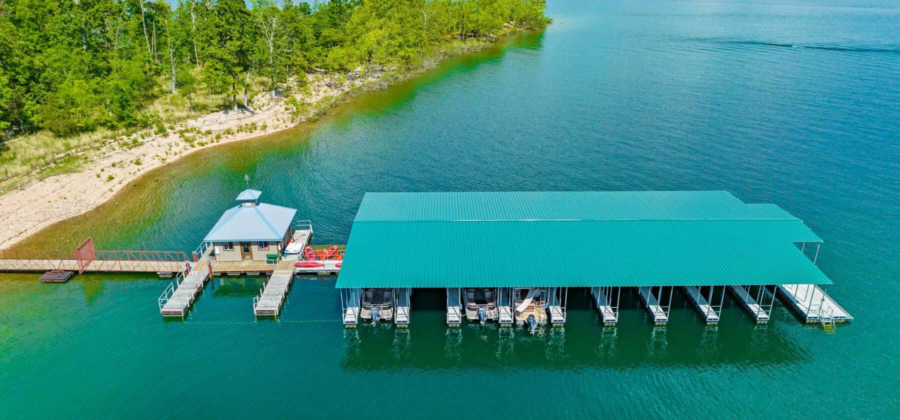 Aerial view of a large boat dock with a teal roof on a lake next to a forested shoreline.