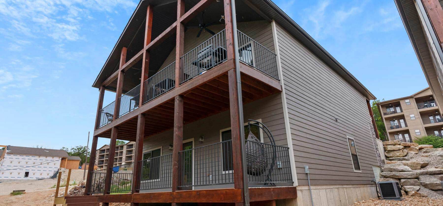 A two-story house with an upper wooden balcony and lower patio against a partly cloudy sky.